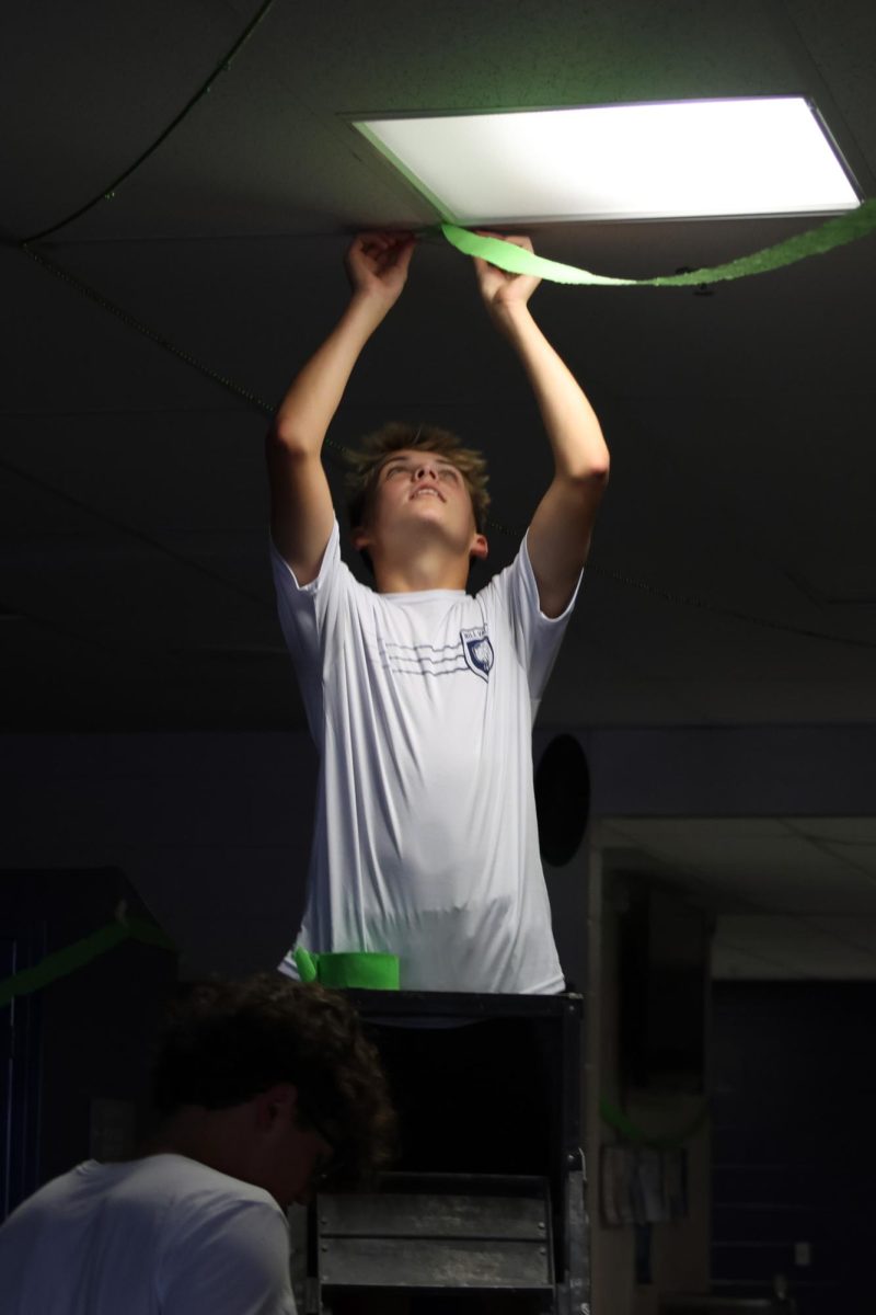 Taping the streamer to the ceiling, sophomore Caden Coup helps decorate the sophomore hallway for homecoming.  