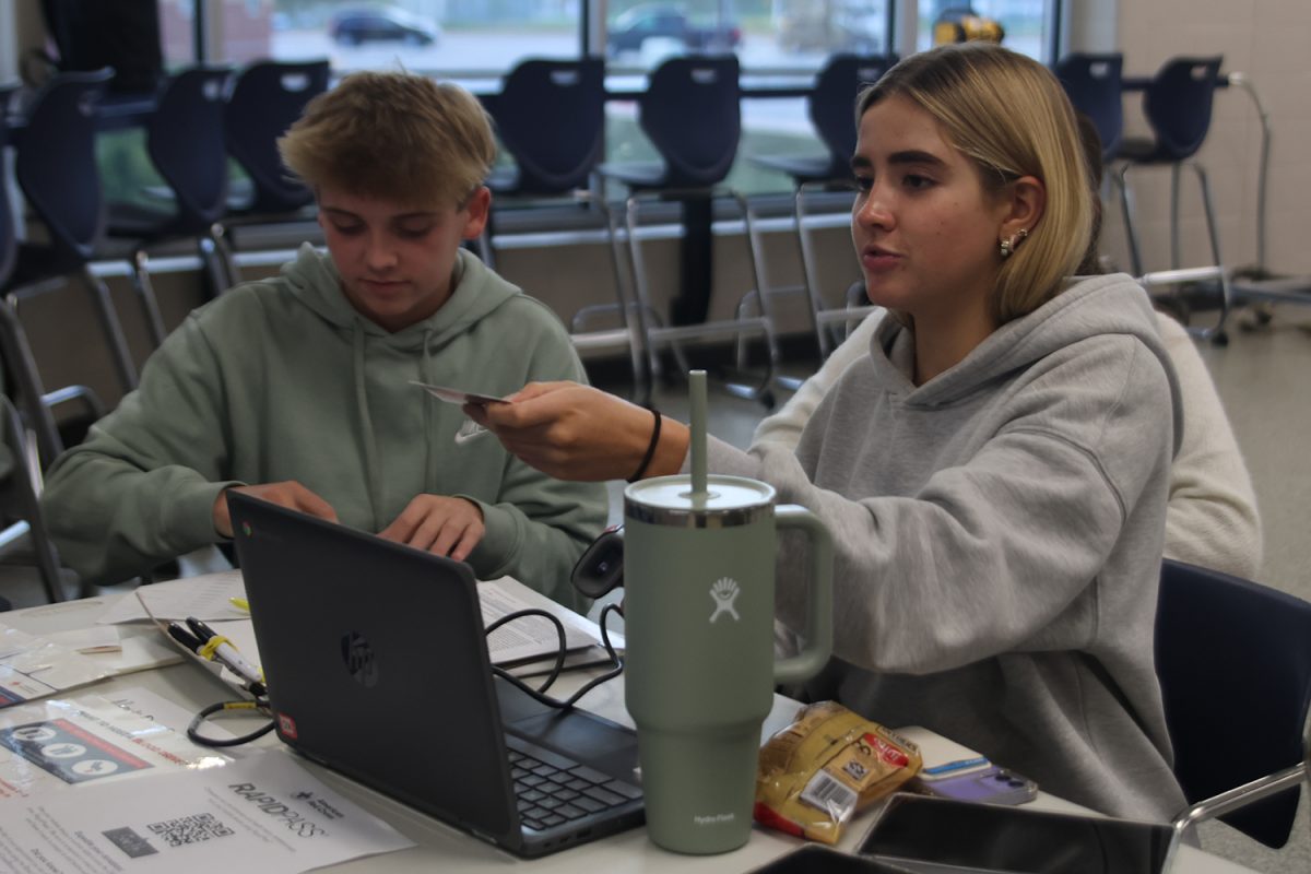 Arm extended, junior Claire Cooper hands ID back to donor while sophomore Caden Coup fills out name tag.
