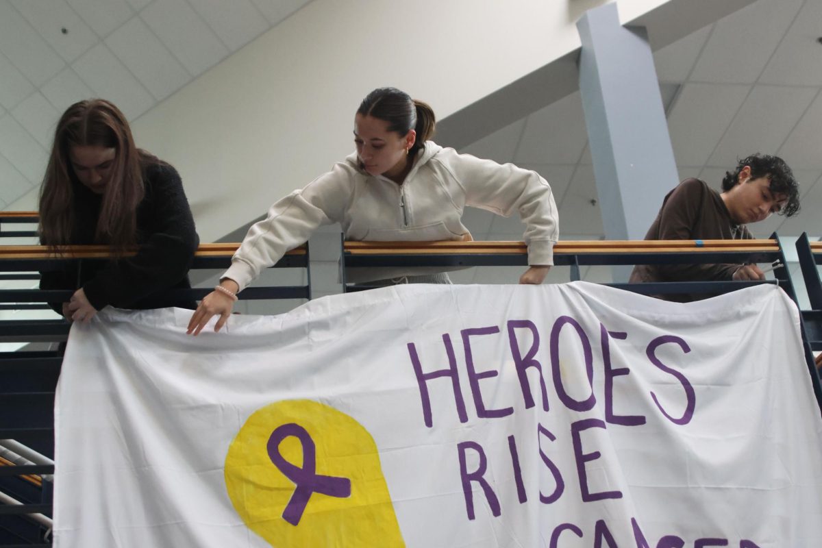 Seniors Emma Ronning, Audri Brizendine, and Maddox Walker tape a large poster across the main staircase during the setup of Relay for Life's Purple Bomb. 